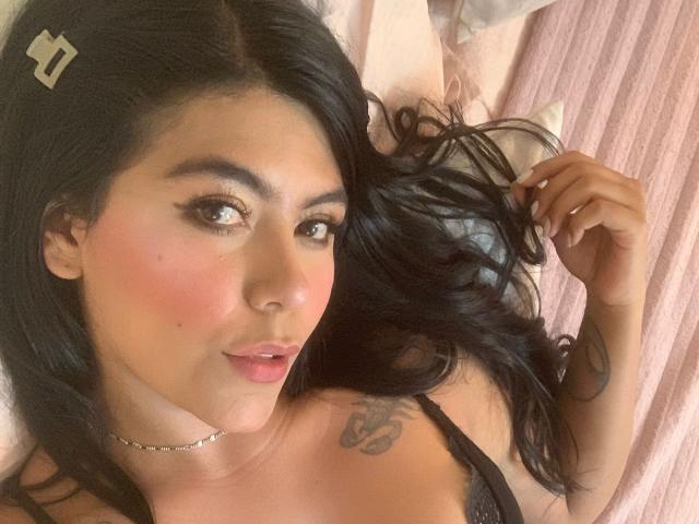 CatalinaReiyna with black hair, pink blush, and a tattoo on her right shoulder, lying down, looking at the camera, holding her hair, wearing a black lace top.