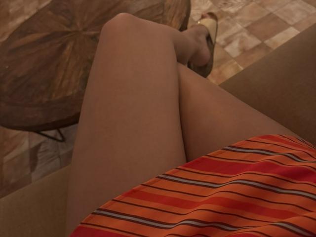 AdharaCooperr's thighs in sheer pantyhose, crossed at the knee, wearing a red and orange striped dress, with brown high heels on a checkered floor.