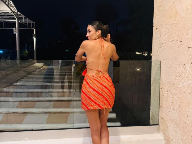AdharaCooperr has a curvy butt in a red, striped halter dress, standing with her back to the camera, looking over her shoulder, on a balcony at night.