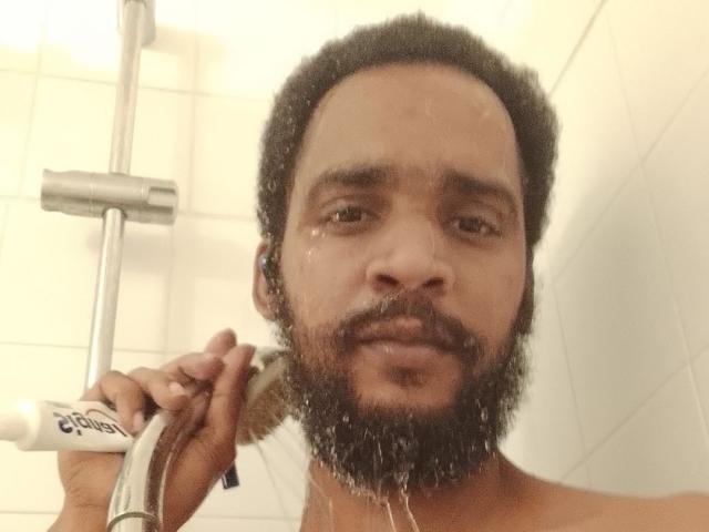 Didikou69 with a dark, curly afro and beard, holding a razor to shave his face in a white-tiled shower, water droplets on his face. Shirtless, looking at the camera.