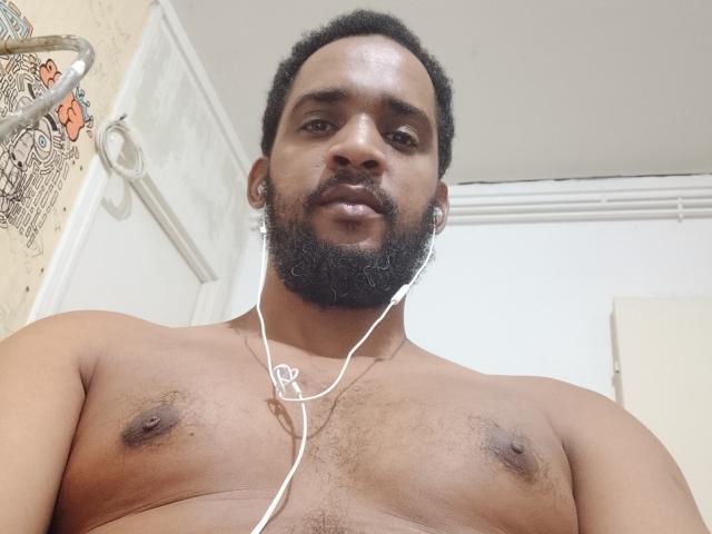 Shirtless Didikou69 with a dark beard, short curly hair, and small nipples, wearing earbuds with white cords, looking down at the camera.