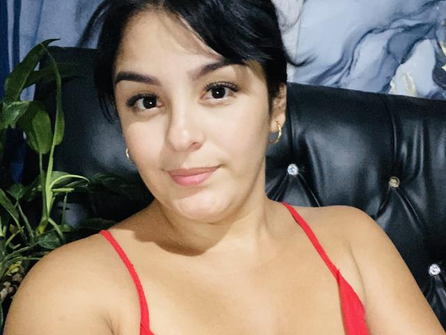 TalianaSquirt with dark hair, brown eyes, light skin, small earrings, and a red top, smiling softly, with a green plant on the left. Boobs visible through top.