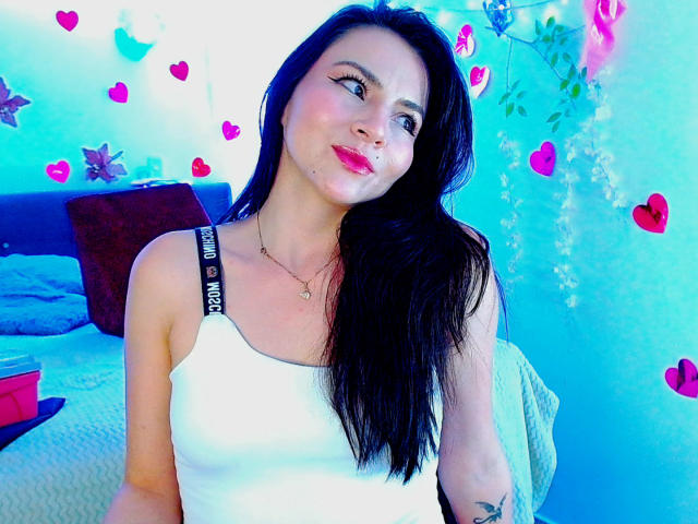 EmelySofia with long black hair, white tank top, pink lipstick, and nose ring, showing off her tits, smiling slightly, in a blue-lit room with heart stickers.