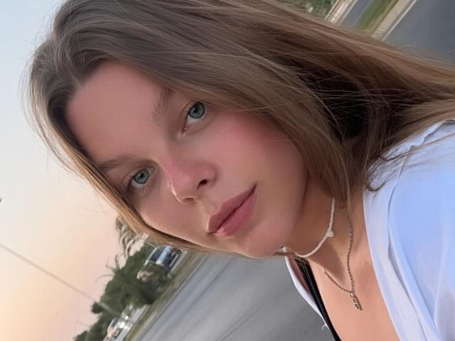 RhonaBells has blue eyes, long brown hair, and pink lips, wearing a white top with a necklace, looking at the camera with a slight smile, in daylight.