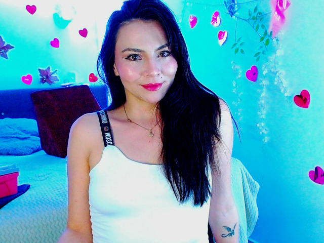 EmelySofia with long black hair, white tank top, pink lipstick, and black bra straps, smiling at the camera, showing a small tattoo on her right arm.
