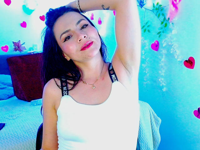 EmelySofia with dark hair, wearing a white tank top, lifting her arm, showing a small tattoo on her side, pink lipstick, and a necklace, in a blue-lit room.