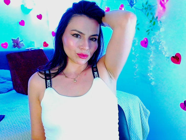 EmelySofia with dark hair, pink lipstick, white tank top, black straps, hand on head, looking at camera, blue and pink heart decorations, subtle makeup, bedroom setting.