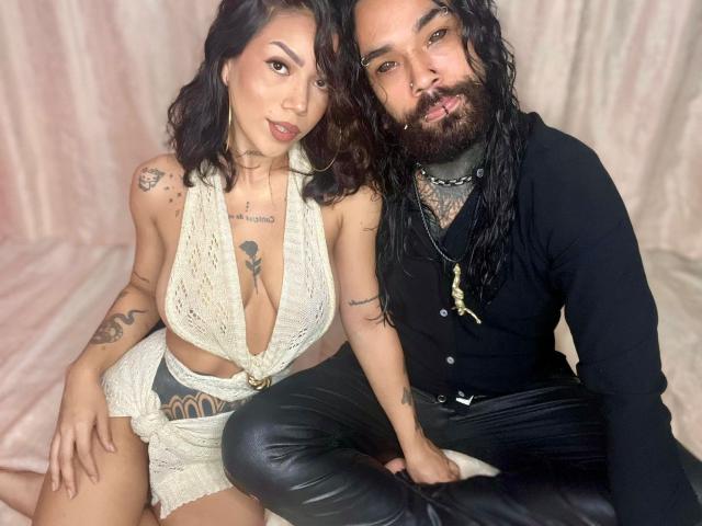 DiogenesYMaria with tits in a white lace halter top and shorts, tattooed, sitting next to a bearded dude in black, leather pants, and tattoos, both looking at the camera.