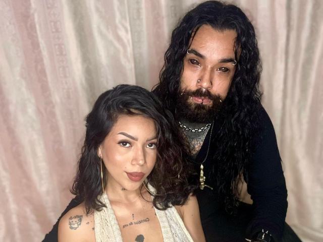 DiogenesYMaria with long curly hair, dark eyes, and tan skin, standing next to a bearded guy with black hair, wearing a black shirt, both looking at the camera.
