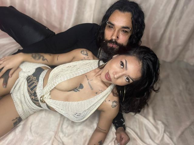 DiogenesYMaria with tattoos and hoop earrings, wearing a white crochet top that reveals her tits, laying on a beige satin sheet, next to a bearded man with black hair.