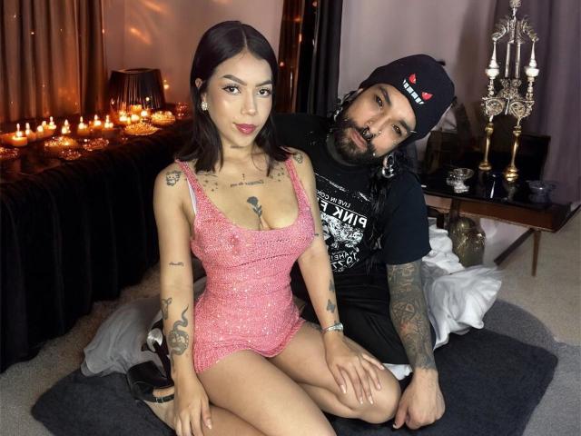 DiogenesYMaria in a pink sparkly dress with small tits, sitting on a rug. Tattooed dude with a beard in a black hat and shirt, kneeling behind her.