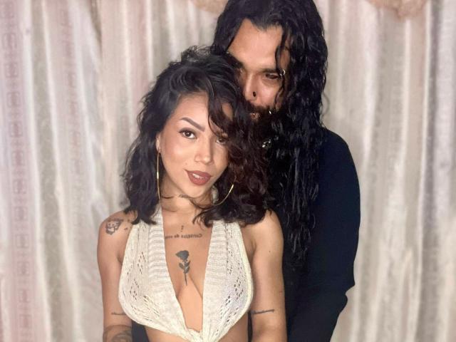DiogenesYMaria with wavy black hair, wearing a white lace halter, showing her tits, tattooed arms, and hoop earrings. Standing behind her is a guy with long black hair.