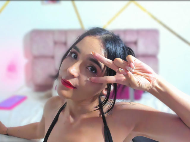 MindyLove, con i capelli neri, il rossetto rosso e un top nero a halter, che fa il segno della pace con la mano destra, mostrando un anello e un tatuaggio sulla spalla sinistra..