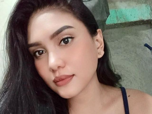 AsianHottie69-hot has smooth, fair skin, dark wavy hair, thick eyebrows, brown eyes, and brown lipstick, wearing a blue top, with a slight smile.
