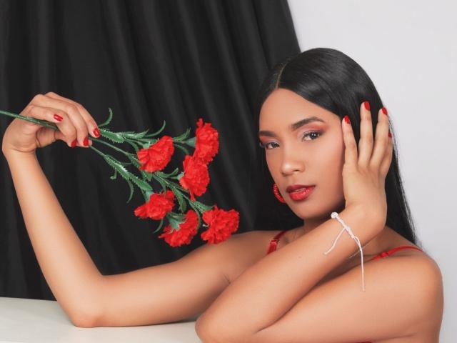 NatLewis with red-painted nails, holding red carnations, wearing red earrings, and a white bracelet, has long black hair, red lipstick, and a sultry expression, against a black curtain.