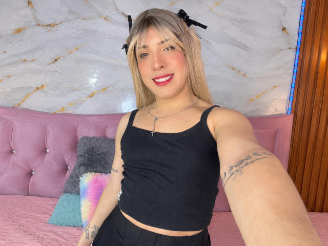AustinCole with blonde hair, black ribbed tank top, black skirt, smiling, red lipstick, tattoos on arm, taking selfie, pink bed, black and pink pillows, wooden wall.
