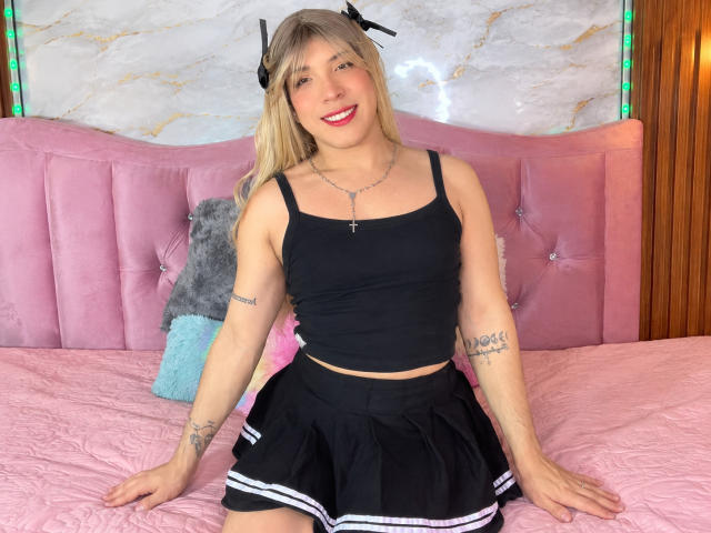 AustinCole with blonde hair, black tank top, and black skirt with white stripes, sitting on pink bed, smiling, tattoos on arms, cross necklace, wooden wall, green lights.