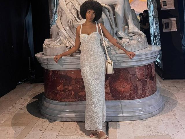 KarlaRicochet è in piedi, indossa un abito bianco di pizzo, che mette in mostra il suo seno piccolo e i suoi fianchi affusolati. Ha una borsa bianca a destra e un grande afro..