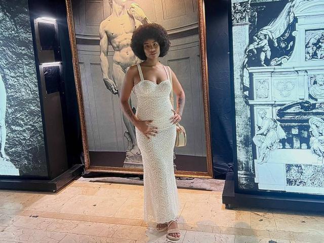 KarlaRicochet está de pie, vestida con un vestido blanco, brillante y ajustado con aberturas, con las manos en las caderas, sosteniendo un bolso pequeño, llevando sandalias beige, con un afro, frente a un cuadro grande enmarcado..