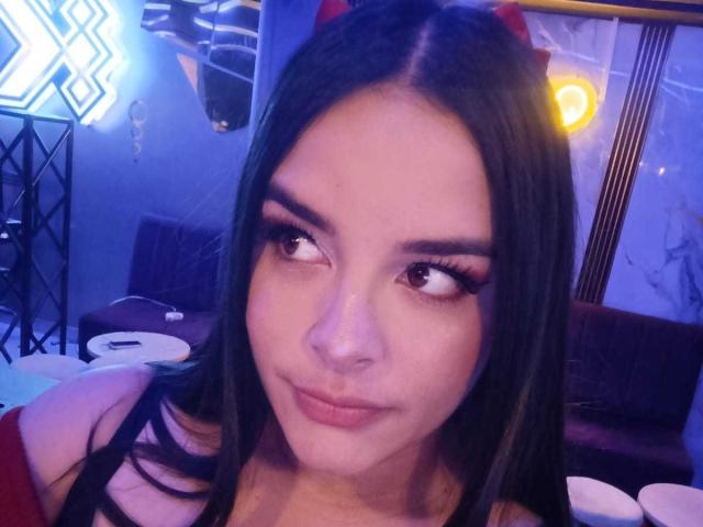 AuroraSantorini has long black hair, dark eyebrows, and wears red devil horns. She's looking to the side with a slight smile, showing her pouty pink lips. Blue lighting.