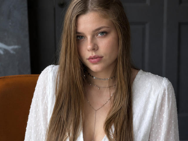 RhonaBells has long brown hair, blue eyes, and is wearing a white, sheer top with a delicate necklace. Her pouty lips and subtle makeup give her a sexy look.