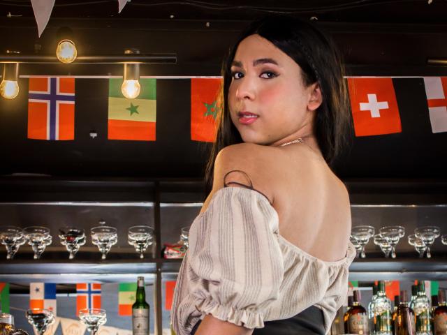 ZoeHotMoretti with dark hair, off-shoulder striped shirt, looking back, exposing her bare shoulders and upper back, standing in front of a bar with glasses and flags.