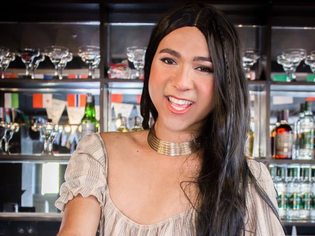 ZoeHotMoretti with long dark hair, smiling with pink lipstick, wearing a beige off-shoulder top and gold necklace, showcasing her cute face and small tits.