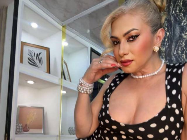 MisseScarleth with blonde hair, wearing a black polka dot dress, pearl necklace, and bracelet, touching her red-painted nails, showing her tits, with a sexy, confident look.