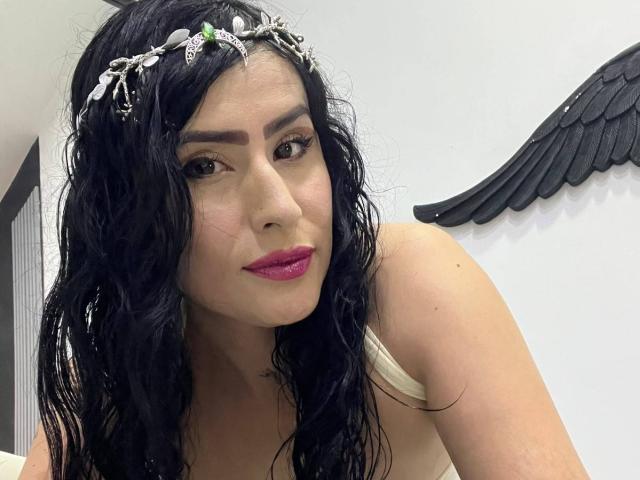 DemiMistral a les cheveux noirs ondulés, un diadème argenté et du rouge à lèvres rose, avec des bretelles blanches sur les épaules, et des décorations sombres en forme d'ailes sur le mur derrière elle..