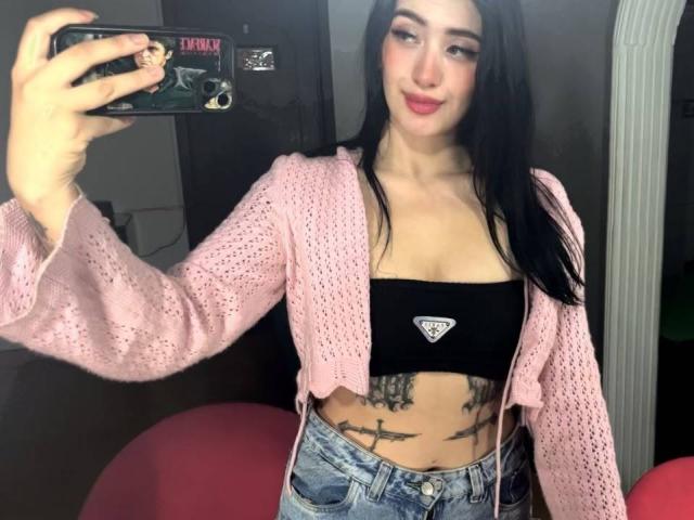TifanyDiaz with black tube top showing small tits, pink cardigan, tattooed belly, blue jeans, holding phone, black case with Tricky design, long black hair, red lipstick, smiling.