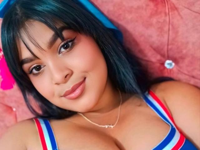 MiaBlakeX tiene el cabello azul oscuro, cejas gruesas y una sonrisa encantadora. Lleva una blusa a rayas rojas, azules y blancas, que deja ver sus grandes senos. Está acostada..