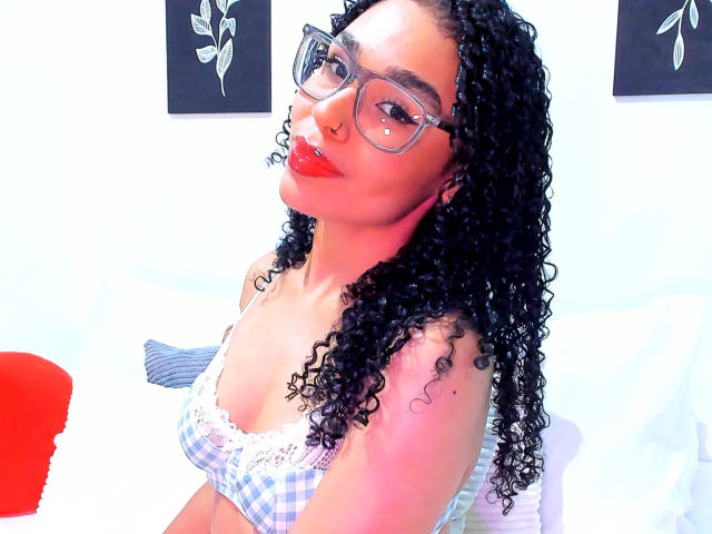 NalaLeroy with curly black hair, glasses, red lipstick, nose ring, checkered bralette, and red lips, sitting on a white bed with a red chair in the background.