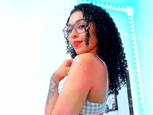 NalaLeroy with curly black hair, glasses, red lipstick, checking her shoulder, showing a tattoo on her right arm, wearing a checkered tank top, smiling.
