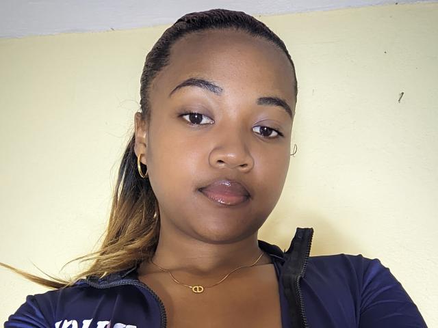 Queenny69 has dark brown skin, medium-sized shiny lips, small hoop earrings, a gold necklace, and is wearing a blue zip-up shirt with her hair in a ponytail.