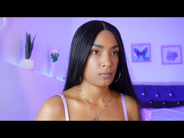 NatLewis has long black hair, brown eyes, and is wearing a pink tank top with a necklace. She has small tits and is looking at the camera with a neutral expression.