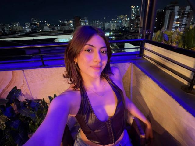 SpencerLe with medium tits in a black leather halter top, blue lighting, brown hair, smiling, on a rooftop at night, city skyline in background.