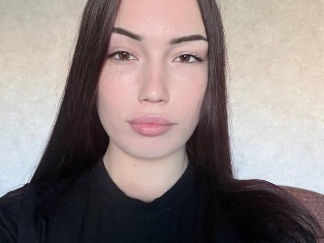Earlie Waldecker tem cabelos longos e castanho-escuros, olhos castanhos e lábios rosados. Ela está vestindo um suéter preto de gola alta, com um rosto neutro, levemente corado, e olhando diretamente para a câmera..