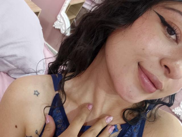 MiiaBennet with wavy black hair, blue lace top, pink nail polish, star tattoo on shoulder, black eyeliner, and a slight smile, holding her chest.