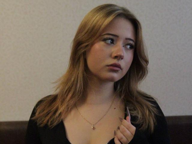 SerafinaDinwiddie with blonde hair, black top, and necklace, looking to the side with a pouty, slightly annoyed expression, holding her shirt collar.
