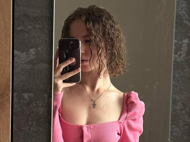 NylaDoore with curly brown hair, pink off-shoulder top, holding a black phone, mirror selfie, necklace with cross pendant, pale skin, subtle smile, visible collarbone, medium-sized tits.