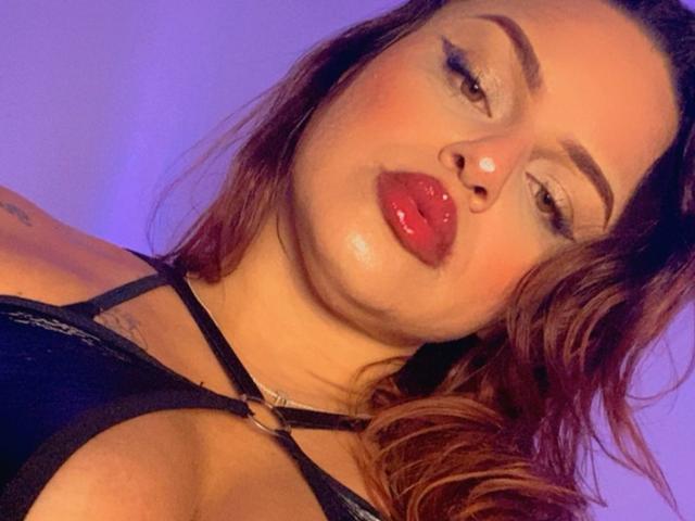 AlmaMilao has puffy red lips, slightly upturned, wearing a black strappy top, with brown wavy hair and subtle tattoos on her shoulder. She's giving a sultry, half-lidded look.