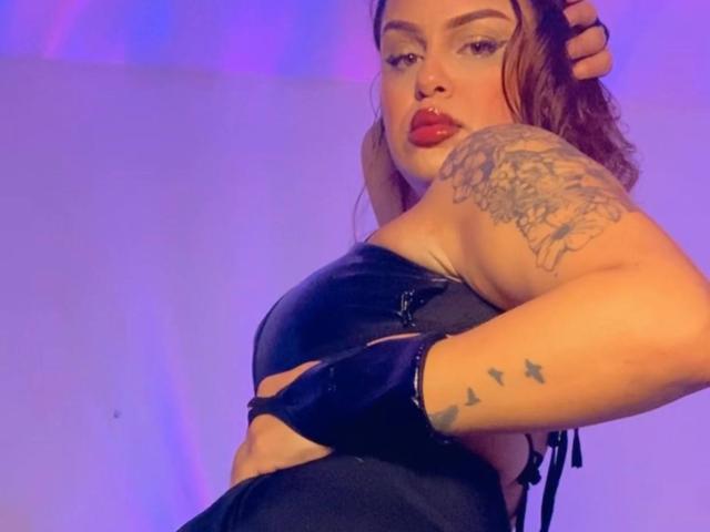 AlmaMilao with red lipstick, black leather bra, tattooed arm, and hand in brown hair, wearing black gloves, looking over shoulder, purple-blue lighting. Boobs, ass, tits visible.