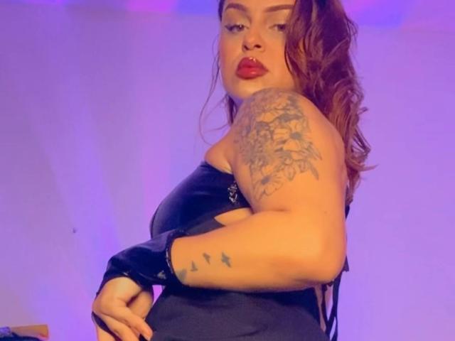 AlmaMilao with red lipstick, black strapless dress, and tattoos on her arm and side, looking over her shoulder, her brown hair in a ponytail, purple lighting.