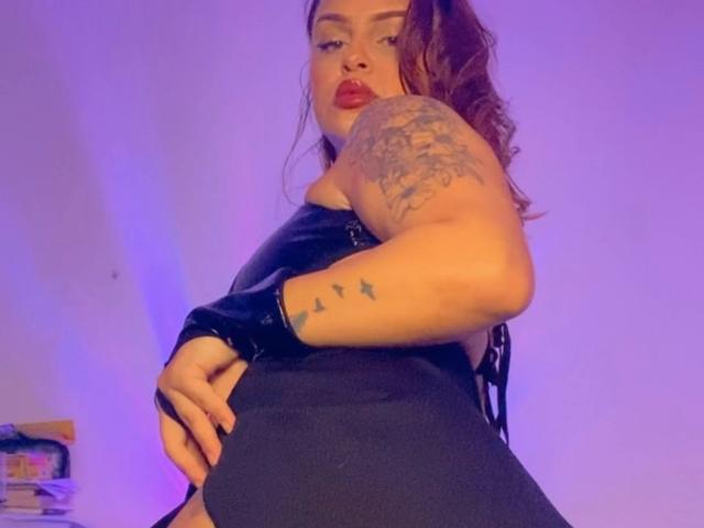 AlmaMilao with red lipstick, black leather top, and black shorts, showing tattoo on arm, raised butt, and wrist bracelet, in purple-lit room. Her ass is prominent.