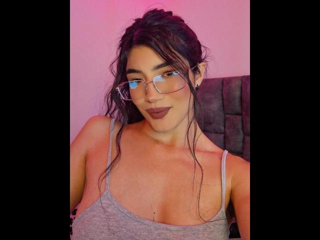 PaulhettJoens with glasses, dark brown wavy hair, and grey tank top, showing her tits and dark lipstick, in pink and purple lighting, looking at the camera.