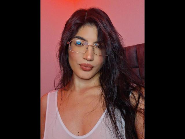 PaulhettJoens with long black hair, wearing glasses, white tank top, and pinkish-red lipstick, showing subtle cleavage and a calm expression. Red light above.