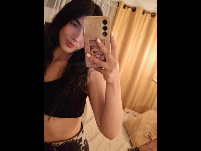 PaulhettJoens with long black hair, wearing a black crop top, taking a mirror selfie with a gold phone case, showing a slim, cute face and small tits.