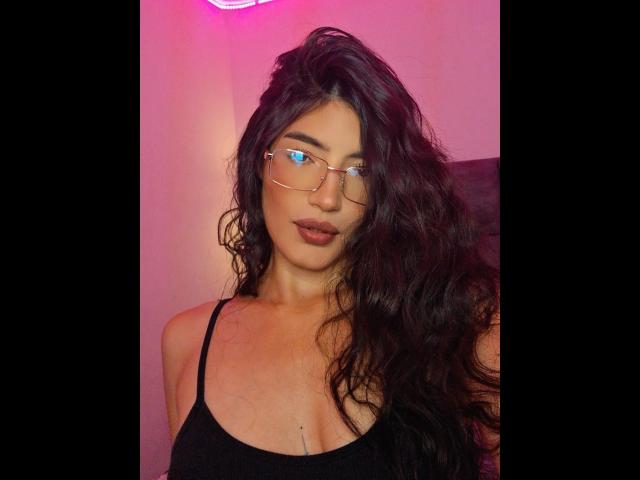 PaulhettJoens has wavy dark brown hair, wearing glasses, a black spaghetti strap top, and has a small chest tattoo below her boobs, with pink neon light in background.