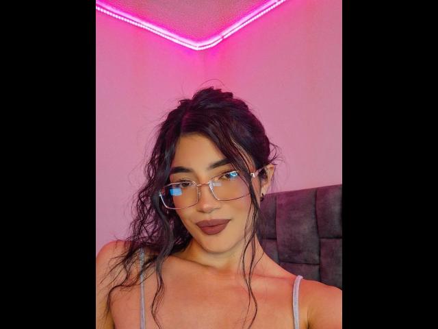 PaulhettJoens with wavy black hair, glasses, and dark lipstick, wearing a light gray top, against pink neon lighting. Boobs visible under top. No makeup on face.
