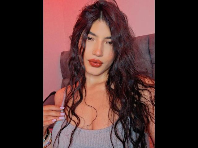 PaulhettJoens with long, wavy black hair, light skin, red lipstick, and a gray tank top, holding a strap, looking at the camera. Boobs visible, hands on strap.